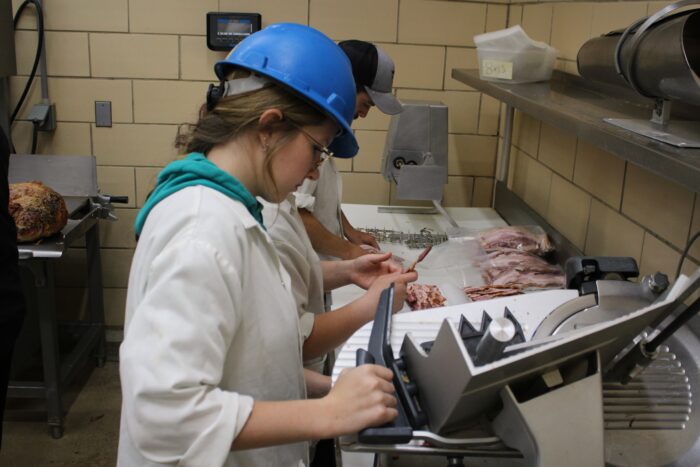 A student wearing a hard hat and a white butcher's coat uses a slicer in a yellow tiled room. Next to her other students can be seen preparing the sliced meat to be vacuum sealed. Behind them a ham hock sits ready to be processed.