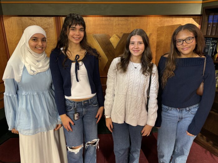 Four high school, young women pose and smile for a photo. The young woman on the far left is wearing a hijab, the other three girls' hair is down. Three young women are wearing jeans and dressy shirts. One young woman is wearing a skirt and blouse.