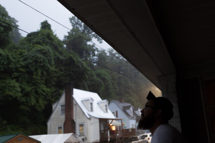 A man standing underneath a building watching rain fall. There are other houses and trees nearby.