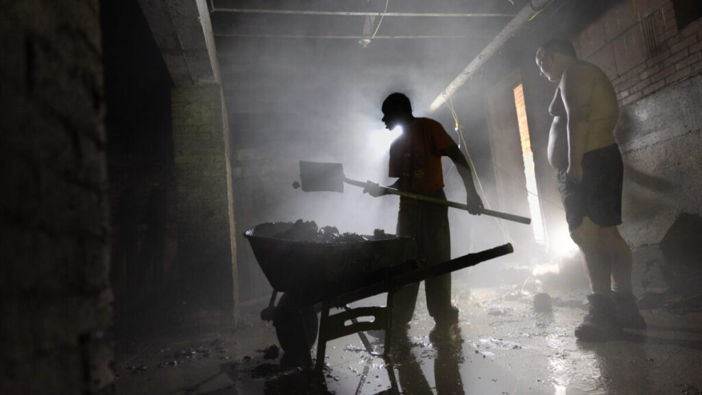 Two men scooping debris with a shovel into a wheelbarrow.
