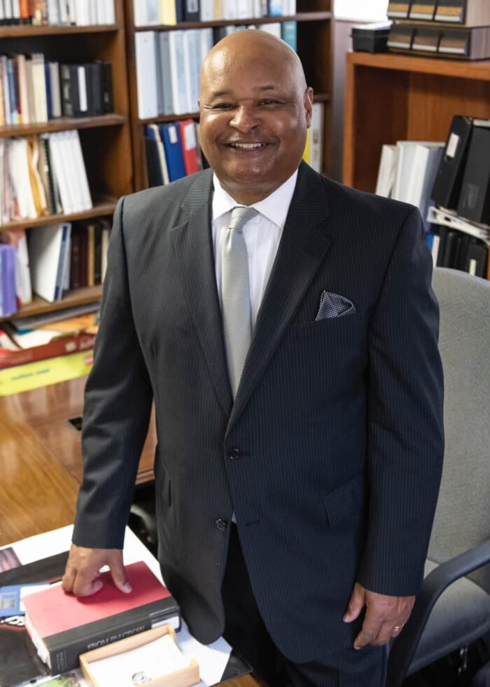 A bald, adult Black man smiles toward the camera. He wears a black suit with light blue tie. He stands in front of bookshelves. His hand rests on a closed book on a desk.