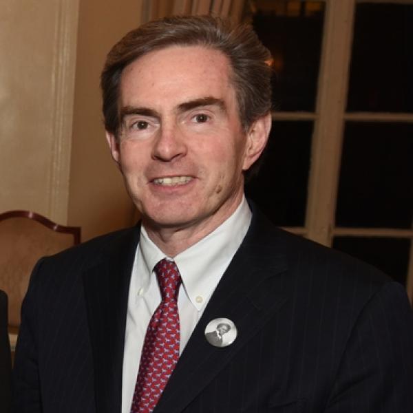A professional headshot of a man with light brown hair. He smiles toward the camera and wears a black suit with a red-checkered tie.