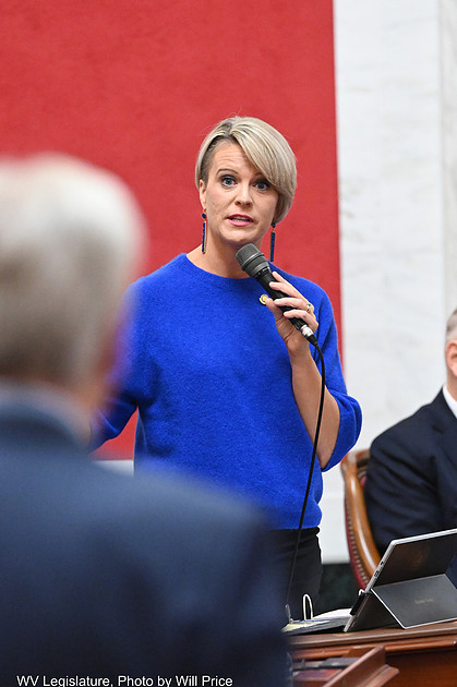 A woman with short blond hair looks toward the camera as an older man with gray hair is blurred looking toward her.