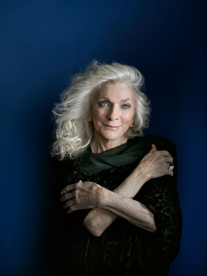 A professional photo of an older woman with long white hair against a dark blue backdrop. She wears a black dressy shirt and crosses her arms against her heart.