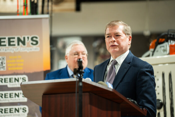 A middle-aged man in a dark suit with white shirt and tie speaks from a podium. Behind him a white haired man in a suit looks on.