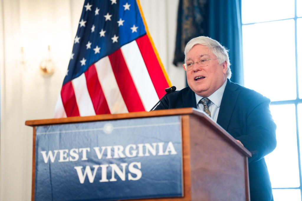 A white-haired man in a navy business suit stands at a podium with the words "West Virginia Wins" across the front. Behind him are an American flag to the left and a West Virginia flag to the right.