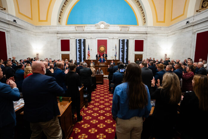 A crowd of professionally dressed people face a man speaking at a podium in a large ornate room