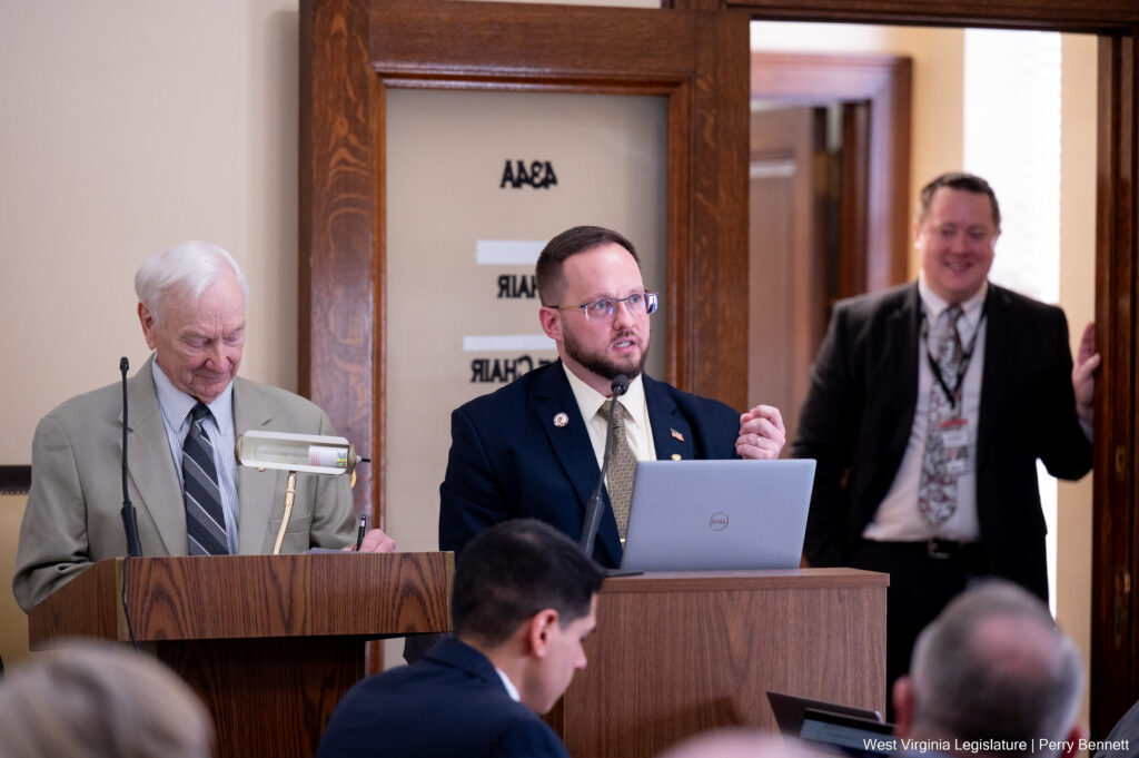 A white-haired man in a light grey suit looks down at a wooden lectern while standing next to a man wearing glasses in a dark blue suit. The spectacled man looks out at a room while speaking and gesturing with one hand over an open laptop computer. He stands in front of an open door with stenciling on a glass panel while a man in a blue suit leans on the door frame.