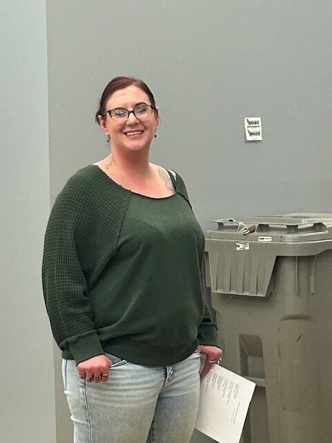 A woman stands next to gray trash cans and looks toward the camera, smiling. She wears a green, long-sleeved shirt and glasses.