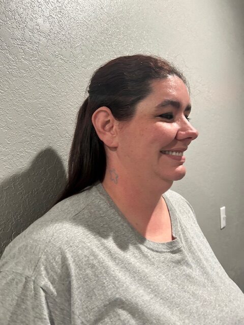 A woman stands against a wall and looks off camera, smiling. She has brown hair and a small star tattoo on her neck.