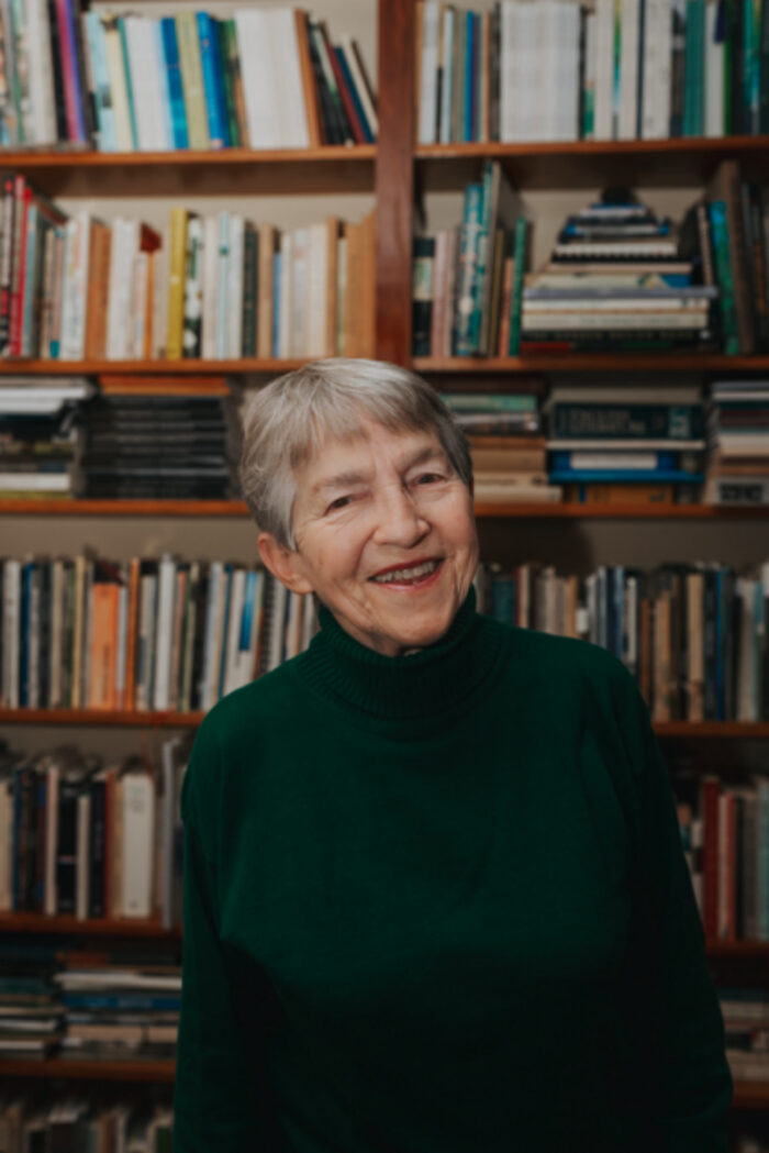An older white woman standing in front of a bookshelf. 