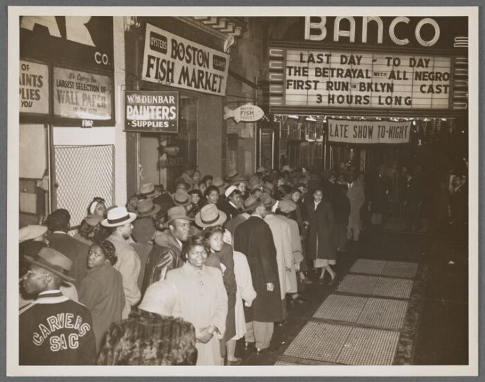 A black and white photo of a crowd of people standing outside a movie theatre.