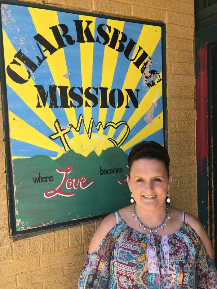 A middle aged woman smiles and looks toward the camera. Behind her is a sign that reads, "Clarksburg Mission, where love becomes..." and the rest of the sign is not shown. The woman is wearing a colorful dress shirt and earrings. Her hair is up in a bun.