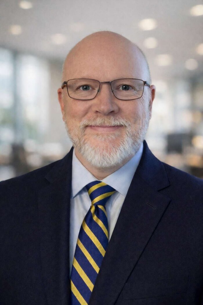 A portrait of a bald man with a white goatee beard wearing glasses. He wears a dark blue suit over a light blue dress shirt and a blue and yellow striped tie.