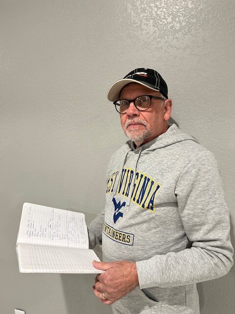 A middle aged man wearing a ball cap and a WVU hoodie looks toward the camera while holding a notebook.