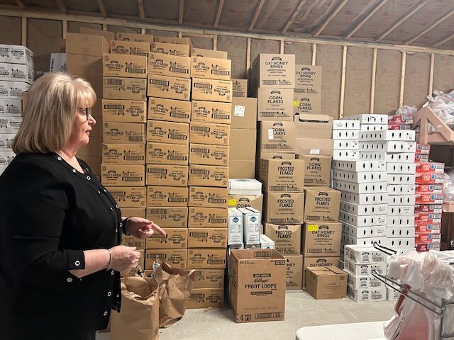 A woman inside a warehouse. She looks off camera. Behind her are boxes stacked almost to the ceiling.