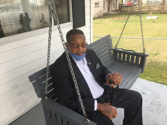 An elderly Black man sits on a porch swing and looks up toward the camera.