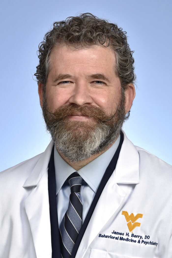 A professional headshot of a doctor. He is middle aged and has curly brown and gray hair, beard, and mustache. His doctors coat reads, "James H. Berry, Behavioral Medicine & Psychiatry."