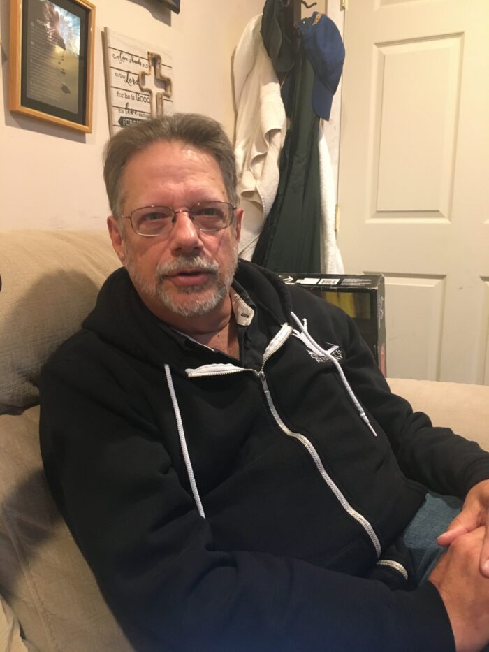 An middle aged man with gray hair and a trimmed gray beard and mustache sits in a chair and speaks to someone. He is wearing glasses, jeans, and a black zip-up hoodie.
