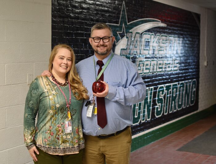 Two middle age people, a man and a woman, pose for a photo smiling. The man holds a red Blenko Glass apple paperweight. They are both dressed business casual.