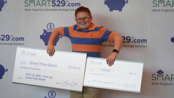 A young boy in a blue and orange striped polo shirt holds up two novelty checks in front of a Smart529 backdrop with piggy bank images