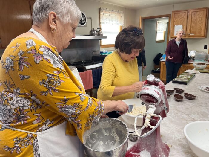 Two women wearing yellow shirts mixing something in a bowl. A woman wearing black is standing farther away. They are in a kitchen. 