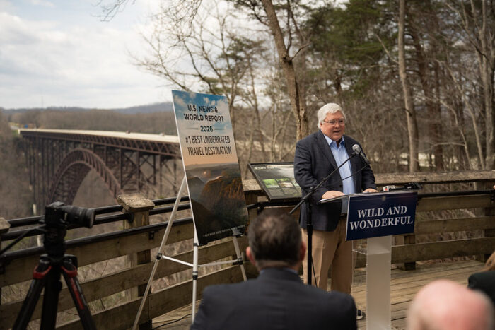 A white-haired man in a navy jacket speaks from a podium overlooking the New River in Fayette County.