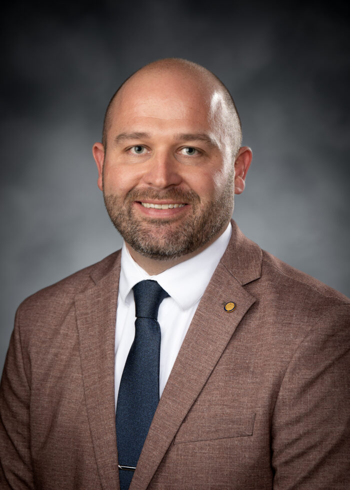 A portrait of a man wearing a light brown suit with a white shirt and a dark blue tie in front of a grey backdrop