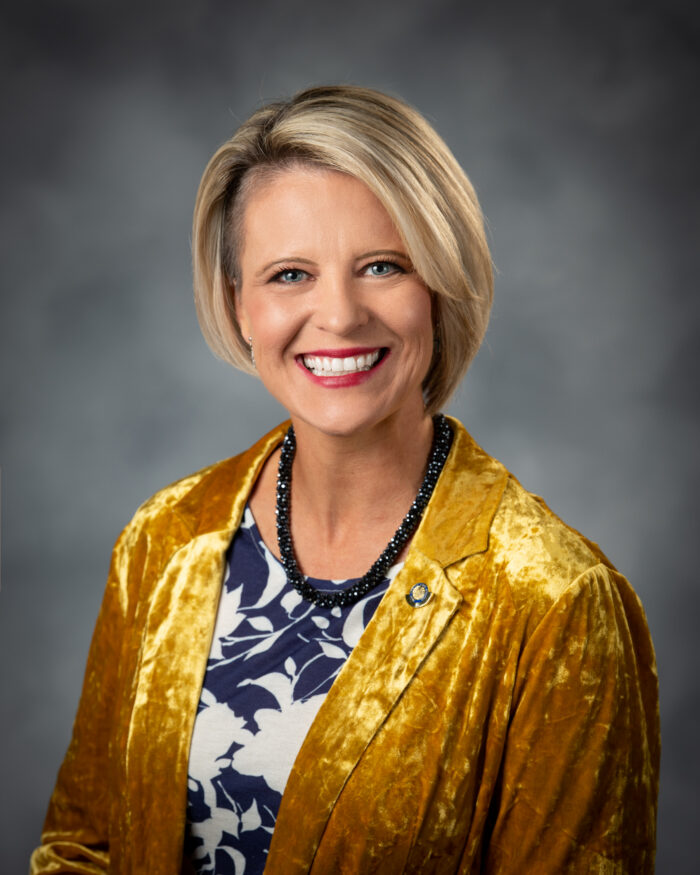 A portrait of a woman wearing a gold blazer over a blue and white blouse in front of a grey backdrop.
