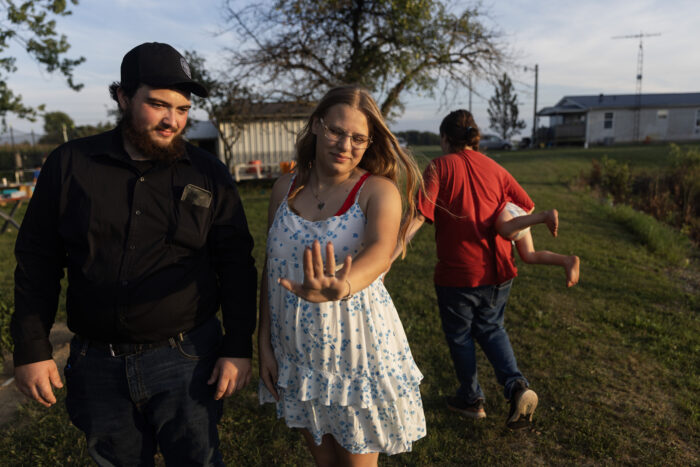 A young white girl holding out her hand to look at an engagement ring. She is standing next to her fiance outside of someone's home. 