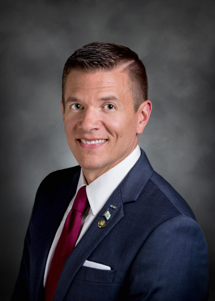 A portrait of a man wearing a dark blue suit with a white shirt and a red tie in front of a grey backdrop