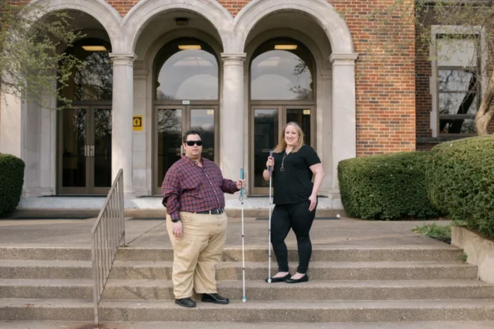 Two adults, a man and a woman, stand on the steps of a higher education institution. They are both holding white canes. The man is wearing sunglasses.