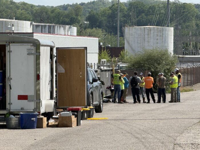 A dozen cleanup workers are seen far from the camera standing together nearby Catalyst Refiners. It's a sunny day. A parked truck is also visible with an open door.
