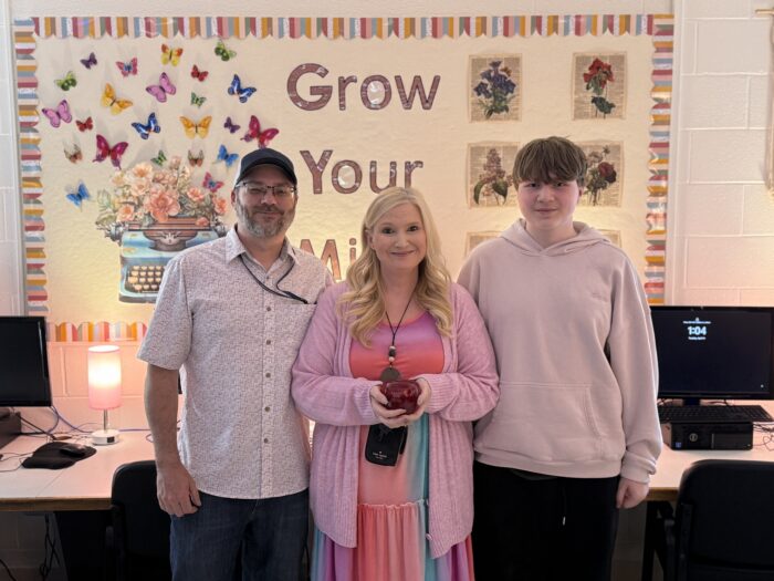 Three people, two adults and one teenager, pose for a photo in a computer lab against a bulletin boards that reads, "Grow Your Mind."