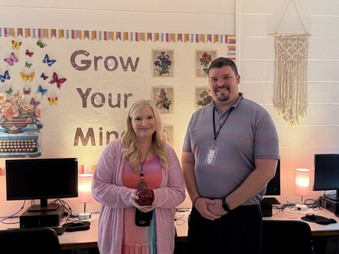 Two adults, a man and a woman, pose for a photo. They are standing in a computer lab against a bulletin board that reads, "Grow Your Mind." She is holding a red, Blenko glass apple paperweight and is wearing mostly pink clothing.