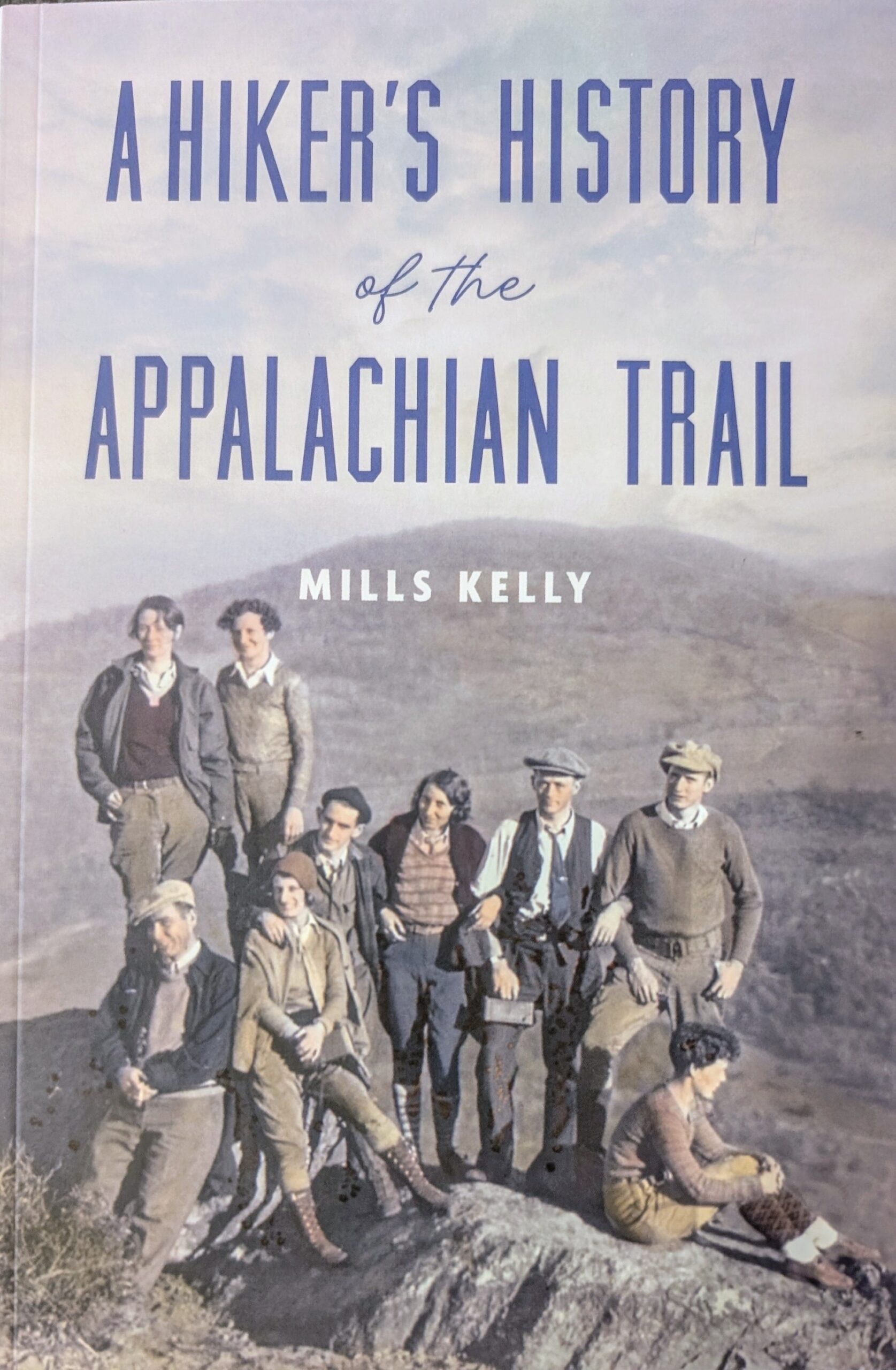 The cover of a book with the title, "A Hiker's History of the Appalachian Trail" by Mills Kelly. There is a photo of nine people posing on the Appalachian trail.