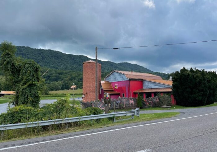 The exterior of Cuz's BBQ. There is a brick silo and a red exterior wall. The restaurant faces a river and mountains. 