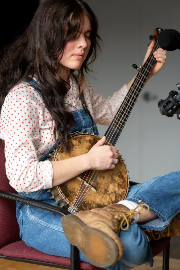 A white woman strumming a banjo. She is wearing blue overalls and a white shirt with orange polkadots. 