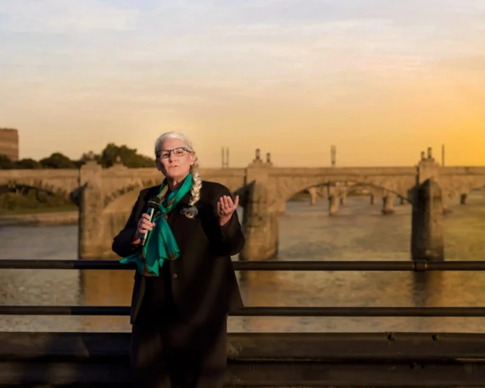 A white woman standing on a bridge in front of a river. She is wearing a green scarf and holding a microphone. 