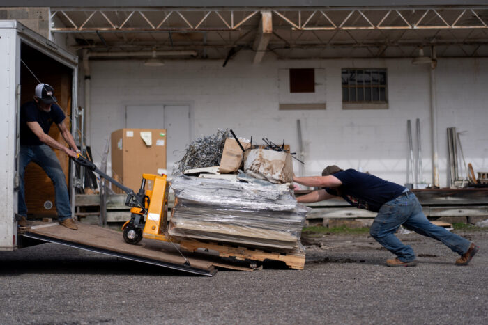 Two men haul materials into the back of a truck. The materials appear to be very heavy.