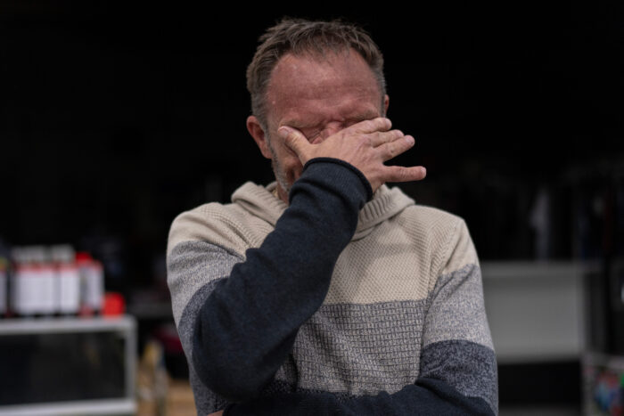 A man in with brown hair presses his right hand into his eyes, crying. He is wearing a long-sleeved sweater.