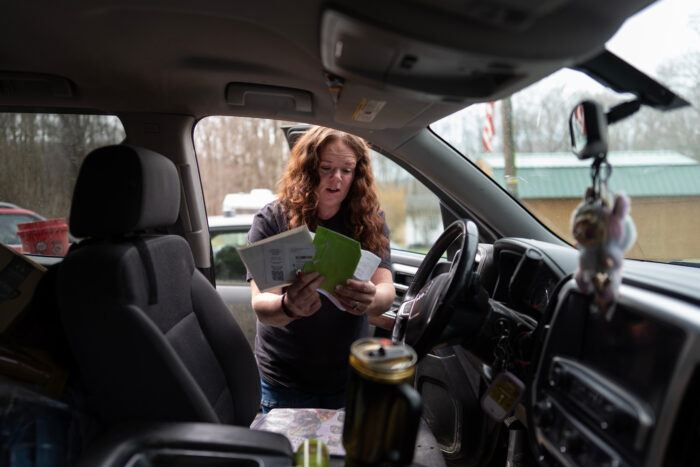 A woman with long red, curly hair leans into a vehicle while holding papers. She appears to be in mid-sentence.
