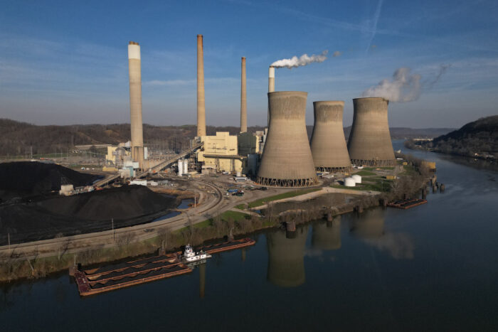 A coal-fired power plan in operation. Smoke billows from two of eight smokestacks against a blue sky. Surrounding the power plant is a river and rolling hills.