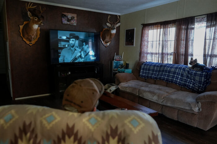 A man in a ball cap sits facing away from the camera in a chair. He watched a black and white film on television. Across from him is a couch. A chihuahua rests on the top of the couch against some windows. On the wall are deer heads on display.