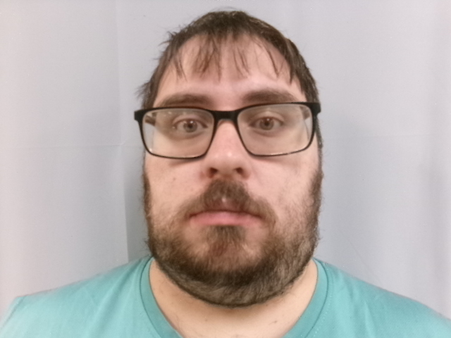Photo shows the head and shoulder of a young man with a scraggly mustache and beard and brown framed glasses wearing a pale green t-shirt against a neutral background.