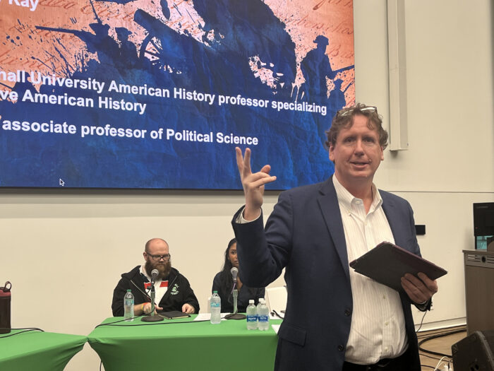 A man in his early 60s stand in front of an audience. Behind him is a panel of speakers. Above the panelists is the lower half of Ken Burns' American Revolution poster.