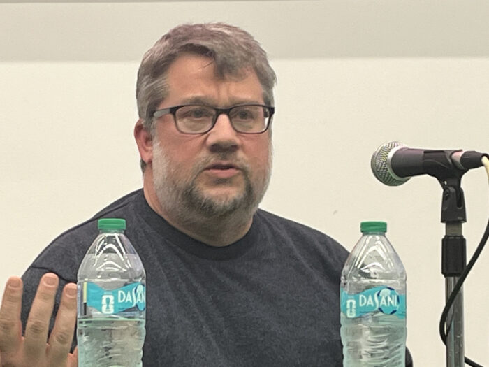 A man with grey hair and a gray beard and mustache wearing a gray t-shirt. He sits in front of a microphone and two water bottles.