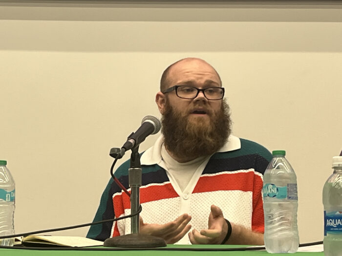 A balding man with a red beard and mustache. He wears glasses and a shirt with blue and red stripes. He sits on a panel.