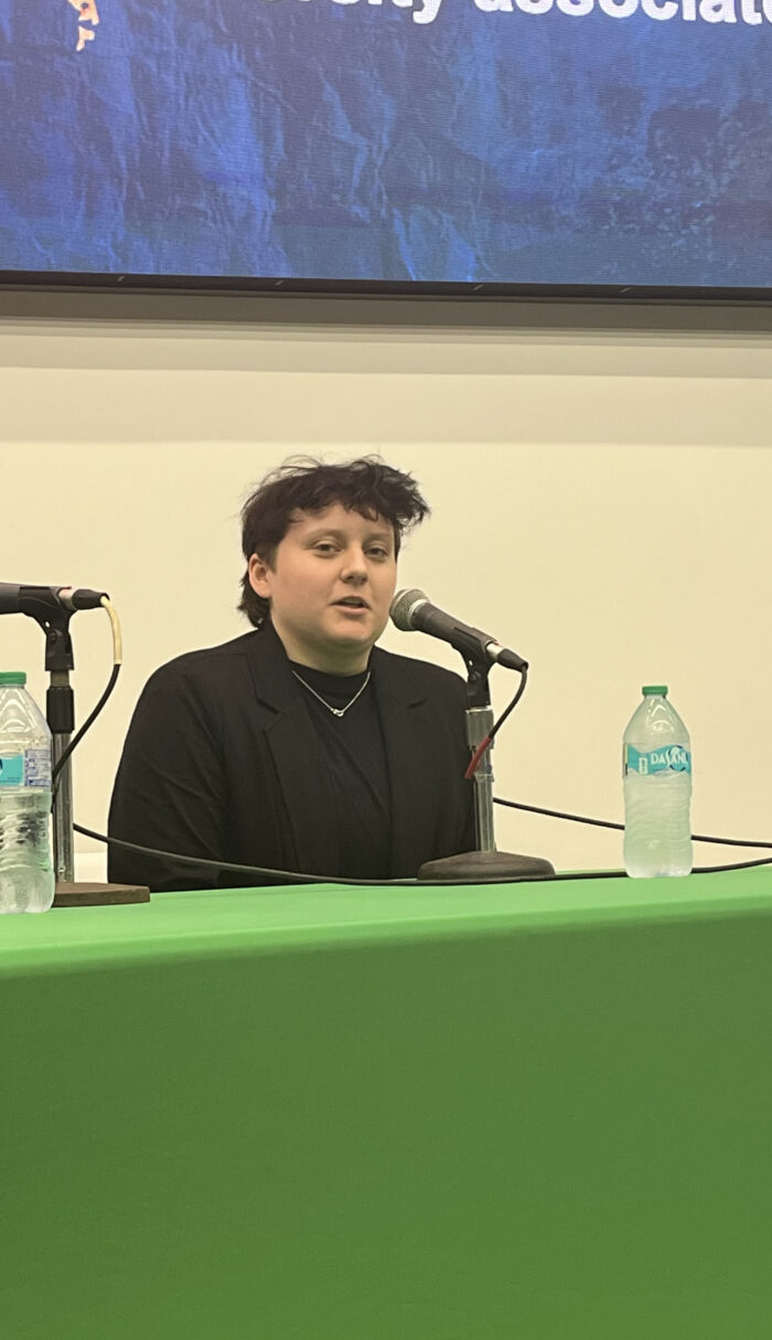 A younger person with shirt dark hair and wearing all black sits on a panel.