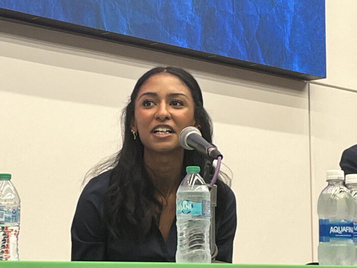 A younger woman with dark hair and wearing a formal, black shirt. She sits on a panel of speakers.